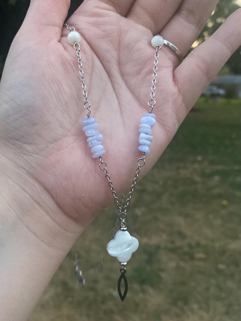May include: A silver chain necklace featuring blue-grey stone chips and white beads. The pendant is a four-leaf clover shape with a dangling black teardrop. The necklace is held in a hand.