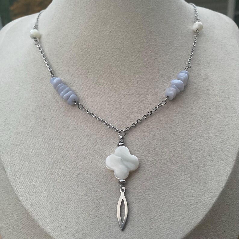May include: A silver-toned necklace featuring a four-leaf clover pendant with a dangling teardrop shape. The necklace has a chain design with light blue and white beads.