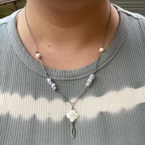 May include: Silver-toned necklace featuring a clover-shaped pendant with a pearl-like bead and a teardrop charm. The necklace has blue-grey stone accents and a delicate chain. The person in the image is wearing a light blue t-shirt.