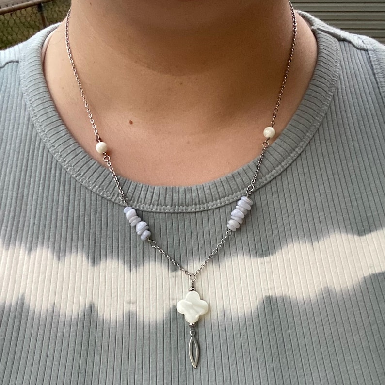 May include: Silver-toned necklace featuring a clover-shaped pendant with a pearl-like bead and a teardrop charm. The necklace has blue-grey stone accents and a delicate chain. The person in the image is wearing a light blue t-shirt.