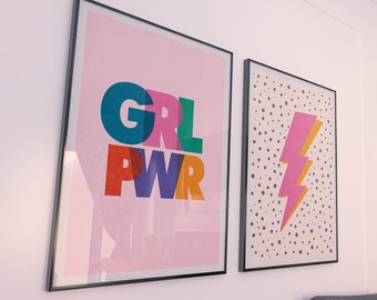 Peut inclure: Deux affiches encadrées sur un mur blanc. L'affiche de gauche a un fond rose avec le texte "GRL PWR" en lettres colorées. L'affiche de droite a un fond blanc avec des pois noirs et un éclair rose et jaune.