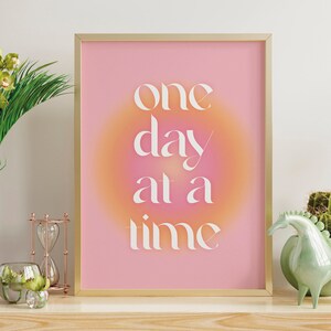 Może przedstawiać: Wzorowany wydruk w ramie z gradientem różowo-pomarańczowym z tekstem "one day at a time" w kolorze białym. Wydruk znajduje się na półce z wazą z zielonymi kwiatami, małym klepsydrą i zieloną ceramiczną figurką konia.