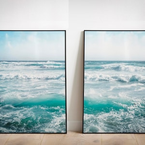 Puede incluir: Dos impresiones artísticas enmarcadas que representan una escena oceánica dinámica. La obra de arte muestra agua turquesa con olas blancas bajo un cielo azul claro. Los marcos son negros y las impresiones se exhiben sobre un suelo de madera.