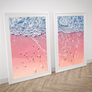 Może przedstawiać: Dwa oprawione wydruki artystyczne przedstawiające widok z lotu ptaka na plażę. Górna część każdego wydruku przedstawia niebieskie fale oceanu, przechodzące w różową plażę z białymi ptakami. Ramy są białe, a grafika jest prezentowana na drewnianej podłodze.