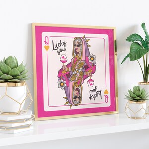 Puede incluir: Una impresi&oacute;n enmarcada de una carta de juego rosa con un dise&ntilde;o de reina de corazones. La reina lleva gafas de sol y un atuendo rosa y dorado. La carta dice "Lucky you" y "Mom's lucky charm" en letras rosa y dorado.