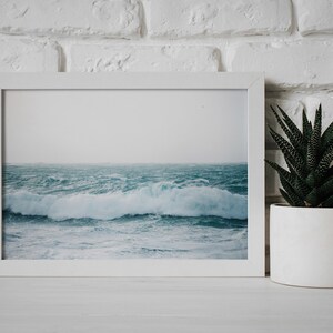 May include: A white framed photograph of a stormy ocean with large waves crashing. The photo is displayed on a white shelf next to a white planter with a green succulent plant.