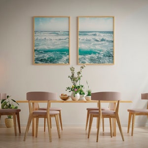 May include: A dining room scene featuring a light wood table and six chairs with pink upholstered seats. Two framed ocean wave prints hang above the table. Decorative plants and pendant lights complete the room's decor.