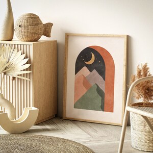 May include: A framed print with a modern abstract design featuring a crescent moon, stars, and mountains in shades of green, orange, and purple. The mountains are framed by a terracotta archway against a dark blue background.