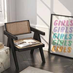 May include: A black cane chair with a woven rattan seat and back, a book on the seat, and a white marble side table with a stack of books on top. A framed print with the word "GIRLS" repeated in different colors hangs on the wall.