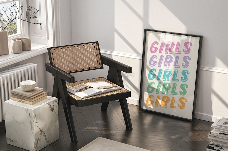 May include: A black cane chair with a woven rattan seat and back, a book on the seat, and a white marble side table with a stack of books on top. A framed print with the word "GIRLS" repeated in different colors hangs on the wall.