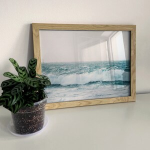 May include: A framed photograph of a seascape with crashing waves. The waves are a deep blue and green color, and the sky is a light blue. The frame is a light wood color.