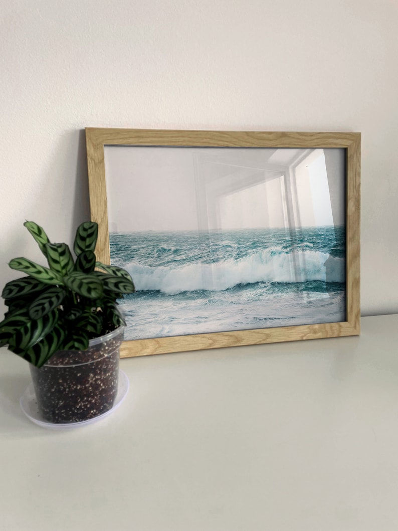 May include: A framed photograph of a seascape with crashing waves. The waves are a deep blue and green color, and the sky is a light blue. The frame is a light wood color.