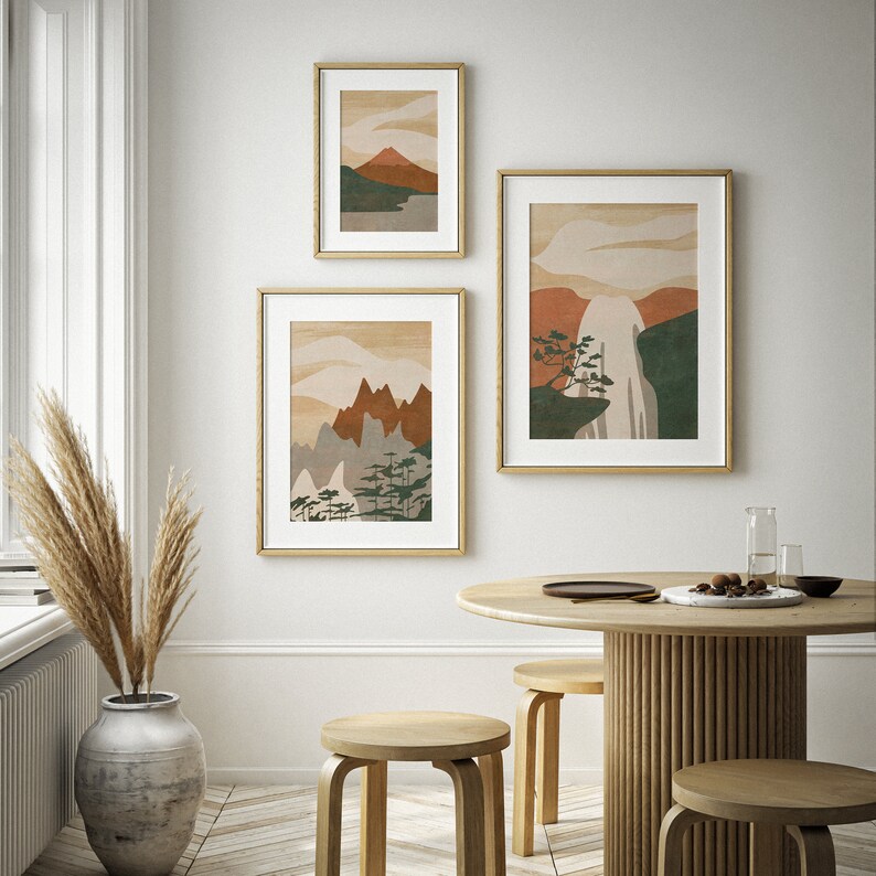 May include: Three framed abstract landscape prints with mountain and waterfall scenes in shades of brown, green, and white, hanging on a white wall above a wooden table with three wooden stools.
