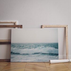 May include: A framed photograph of a stormy ocean scene with large waves crashing against the shore. The photograph is displayed on a wooden floor with a white wall in the background.