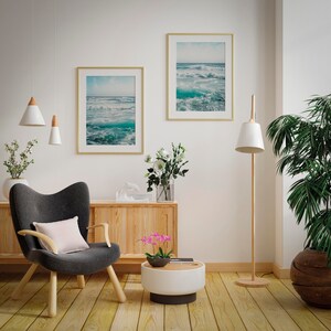 May include: Interior design featuring two framed ocean wave prints with gold frames. A gray armchair with a pink pillow sits near a wooden cabinet and a white and wood side table. A floor lamp and a potted plant complete the room.