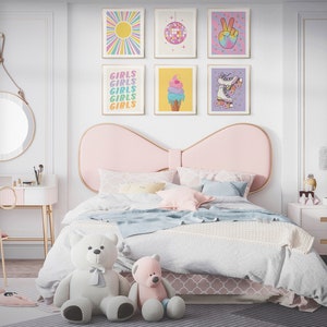 May include: A pink and white bedroom with a bed featuring a large pink bow headboard. The bed has a white and blue blanket and a white and pink patterned bed skirt. There are several colorful framed prints on the wall above the bed, including one with the word "Girls" repeated several times. There is a white vanity with a gold metal frame and a pink bow-shaped chair. A white nightstand with gold accents is next to the bed.