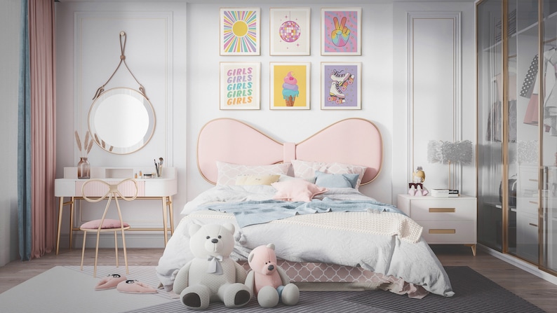 May include: A pink and white bedroom with a bed featuring a large pink bow headboard. The bed has a white and blue blanket and a white and pink patterned bed skirt. There are several colorful framed prints on the wall above the bed, including one with the word "Girls" repeated several times. There is a white vanity with a gold metal frame and a pink bow-shaped chair. A white nightstand with gold accents is next to the bed.