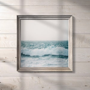 May include: A framed photograph of a stormy ocean scene with large waves crashing against the shore. The photo is in a silver frame and is hanging on a white wood wall.