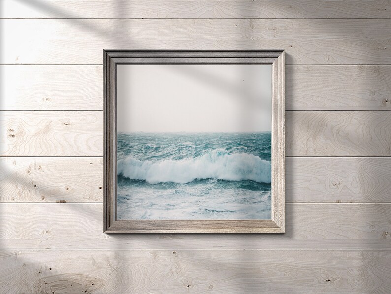 May include: A framed photograph of a stormy ocean scene with large waves crashing against the shore. The photo is in a silver frame and is hanging on a white wood wall.
