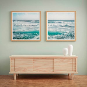 May include: Two framed ocean wave prints with light wood frames hang above a light wood cabinet. Two white vases sit on the cabinet. The room has light green walls and a wood floor.