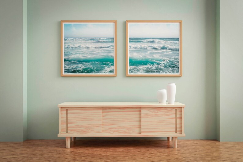 May include: Two framed ocean wave prints with light wood frames hang above a light wood cabinet. Two white vases sit on the cabinet. The room has light green walls and a wood floor.
