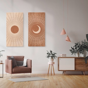 May include: Two minimalist wall art prints with a sun and moon design in shades of brown and beige. The prints are hung on a white wall in a living room setting with a brown armchair, a side table, and plants.
