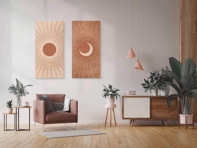 May include: Two minimalist wall art prints with a sun and moon design in shades of brown and beige. The prints are hung on a white wall in a living room setting with a brown armchair, a side table, and plants.