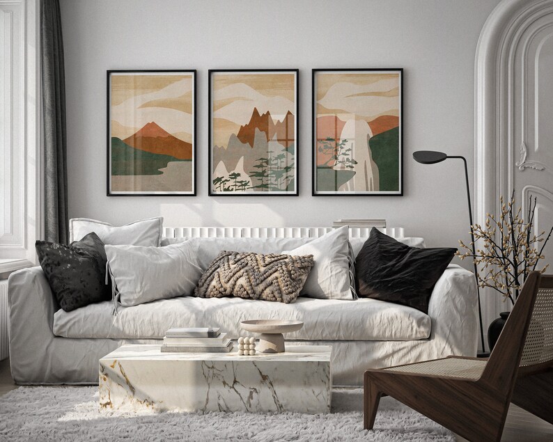 May include: Three abstract landscape prints in black frames hang above a white sofa with black and white throw pillows. A wooden chair with a woven seat sits in the corner of the room.