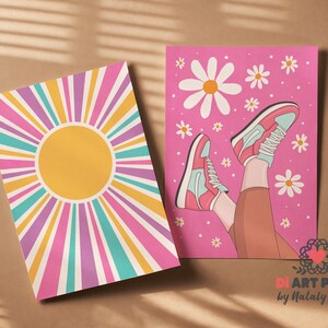 May include: Two art prints. One features a yellow sunburst design with pink, purple, teal, and yellow rays. The other print is pink with a drawing of legs wearing pink and teal sneakers, surrounded by white daisies.