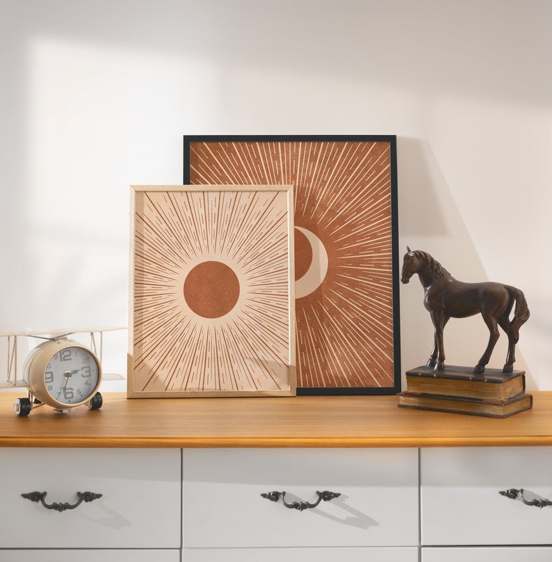May include: Two framed prints with abstract sun and moon designs in shades of brown and beige, displayed on a white dresser with a bronze horse figurine and a vintage clock.