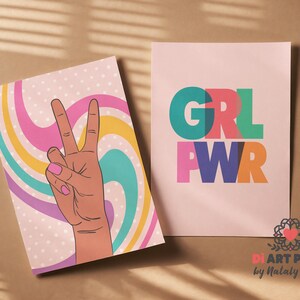 May include: Two art prints. One features a hand making a peace sign with a retro swirl background. The other print says "GRL PWR" in colorful block letters. Both prints are on a brown surface.