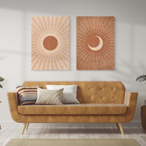 May include: Two minimalist art prints with a sun and moon design. The prints are in a warm terracotta color with a white background. The prints are hanging on a white wall above a brown leather sofa.