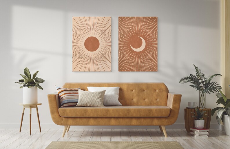 May include: Two minimalist art prints with a sun and moon design. The prints are in a warm terracotta color with a white background. The prints are hanging on a white wall above a brown leather sofa.