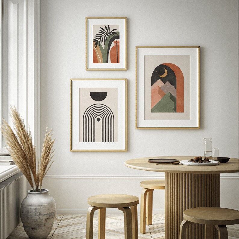 May include: Three abstract art prints with gold frames. The prints feature geometric shapes, a crescent moon, and a stylised palm tree. The prints are hung on a white wall above a wooden table with three stools.