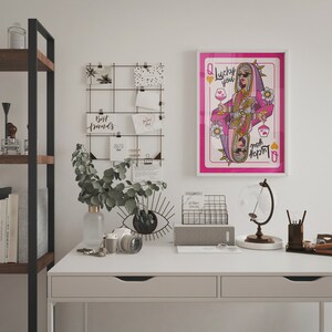 Puede incluir: Un escritorio blanco con dos cajones, una estanter&iacute;a de metal negro, un globo terr&aacute;queo, un calendario, una planta, una c&aacute;mara, un jarr&oacute;n y una impresi&oacute;n enmarcada de una carta de juego de la reina de corazones con el texto "Lucky You" en ella.