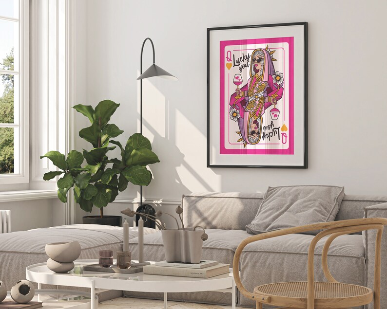 Puede incluir: Una ilustraci&oacute;n enmarcada de una carta de la reina de corazones con una mujer con gafas de sol y un atuendo rosa y dorado sosteniendo una copa de vino. La carta tiene el texto "Lucky you" y "Queen of Hearts".