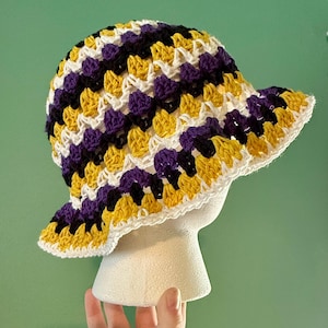 May include: A crochet bucket hat featuring a repeating pattern of yellow, purple, and white yarn. The hat has a rounded crown and a wide, slightly flared brim, constructed with a textured stitch pattern that creates a dense, layered appearance.