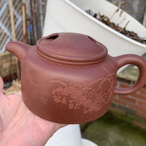 May include: A reddish-brown teapot with a lid and a handle. The teapot has a floral design carved into the side.
