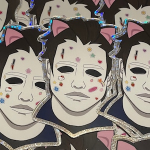 May include: A stack of colorful stickers featuring a cartoon character with a white mask and pink cat ears. The character has various colorful stickers on their face, including hearts, stars, and a rainbow.