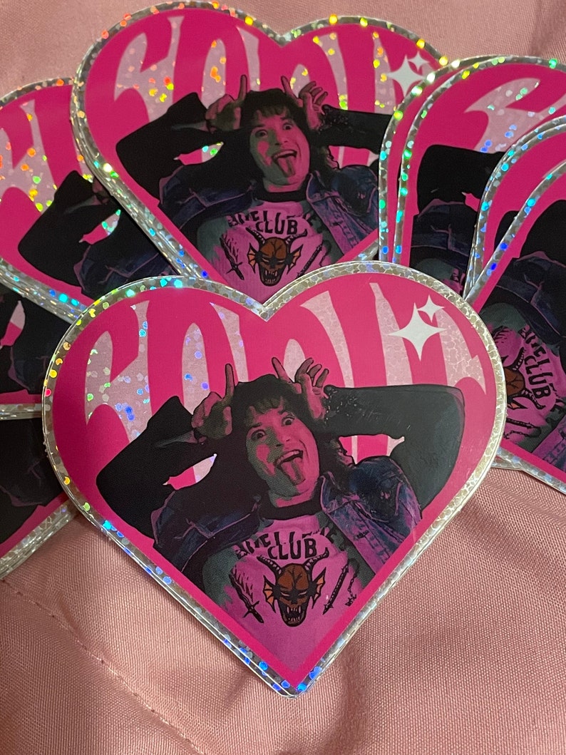 Pode incluir: Adesivo rosa em forma de cora&ccedil;&atilde;o com acabamento hologr&aacute;fico que apresenta uma pessoa com a l&iacute;ngua de fora fazendo uma careta engra&ccedil;ada. A pessoa est&aacute; vestindo uma jaqueta preta, uma camiseta rosa com um gr&aacute;fico de um monstro e as palavras "Hell Club", e um colar preto. O adesivo tem uma borda branca.