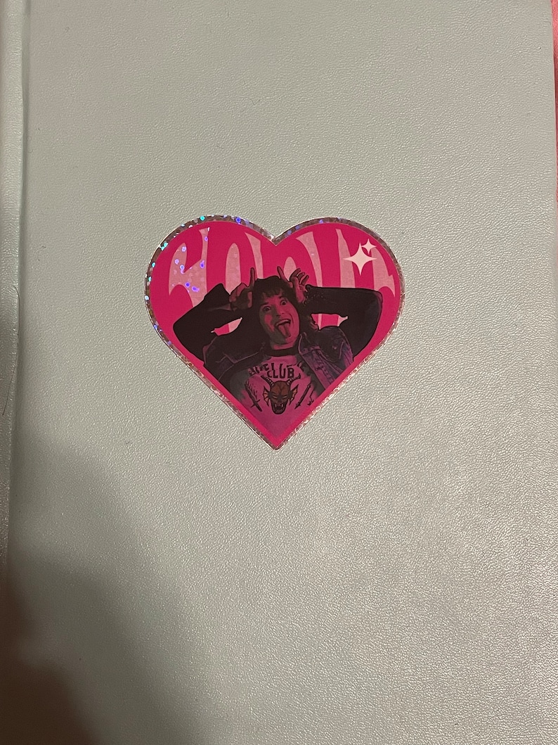 Pode incluir: Um adesivo rosa em forma de cora&ccedil;&atilde;o com acabamento brilhante apresenta uma mulher com cabelo escuro e uma camiseta branca e preta com o texto "Hell Club" e um gr&aacute;fico de lobo. A mulher est&aacute; mostrando a l&iacute;ngua e fazendo uma careta engra&ccedil;ada.