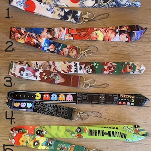 May include: Five colorful lanyards with different cartoon characters and designs. The lanyards are made of fabric and have a black metal clip at the end. The designs include a Japanese wave pattern, a video game character, a cartoon dog, a Pac-Man game design, and a cartoon Martian.