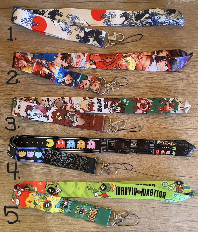 May include: Five colorful lanyards with different cartoon characters and designs. The lanyards are made of fabric and have a black metal clip at the end. The designs include a Japanese wave pattern, a video game character, a cartoon dog, a Pac-Man game design, and a cartoon Martian.