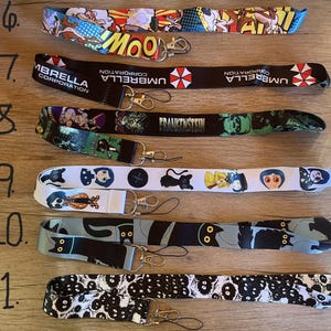 May include: Eleven different lanyard designs featuring various characters and logos. The lanyards are white, black, blue, and red with various graphic designs. The lanyards are numbered 6 through 11.