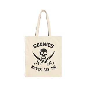 May include: A white canvas tote bag with a black skull and crossbones design. The text "Goonies Never Say Die" is printed below the skull.