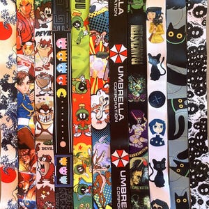 May include: A collection of colorful lanyards with various cartoon and video game characters, including Pac-Man, a black cat, and a character from the video game Street Fighter. The lanyards also feature the text "UMBRELLA CORPORATION" and "FRANKENSTEIN".