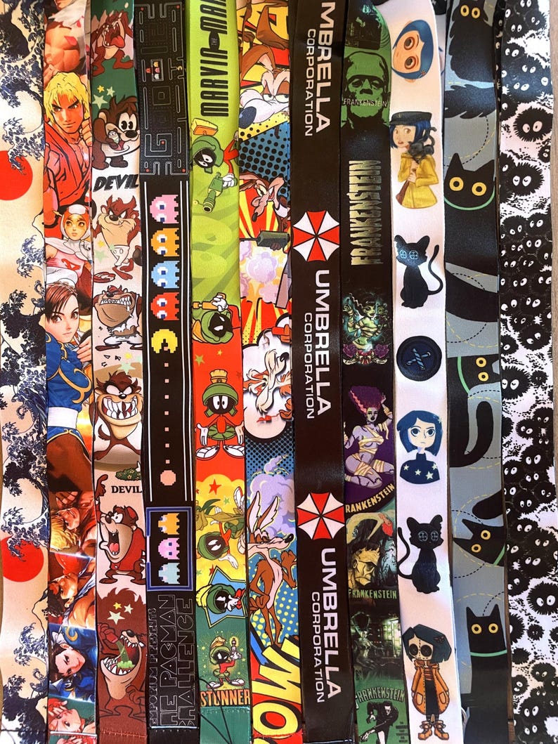 May include: A collection of colorful lanyards with various cartoon and video game characters, including Pac-Man, a black cat, and a character from the video game Street Fighter. The lanyards also feature the text "UMBRELLA CORPORATION" and "FRANKENSTEIN".