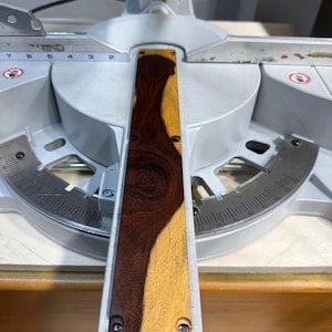 May include: A rectangular wooden insert with a dark brown and light yellow wood grain pattern, featuring four small circular screw holes at the corners, positioned on a silver metal miter saw base with a visible imperial measuring scale.