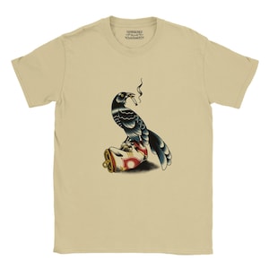 Peut inclure: Un t-shirt beige à manches courtes orné d'un motif représentant un corbeau noir perché sur une canette de soda écrasée, tenant une cigarette allumée dans son bec. L'étiquette intérieure du col porte le texte "CHUPACABRA" et "WhatsGoodAFX".