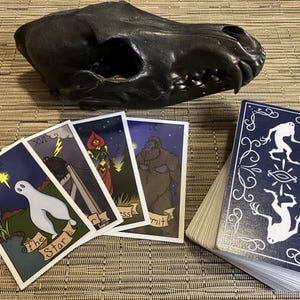 Peut inclure: Un jeu de cartes de tarot avec des illustrations d'un fant&ocirc;me, d'une femme en robe rouge, d'une grande cr&eacute;ature poilue et d'une cr&eacute;ature avec un troisi&egrave;me &oelig;il. Les cartes sont dispos&eacute;es sur un tapis tiss&eacute; avec un cr&acirc;ne d'animal noir en arri&egrave;re-plan.